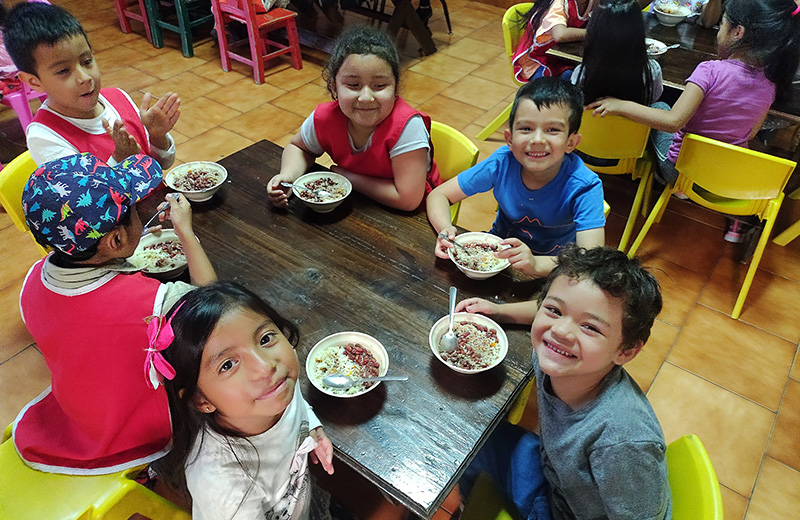 A group of children gathered around a table, enjoying a meal together with plates of food in front of them.