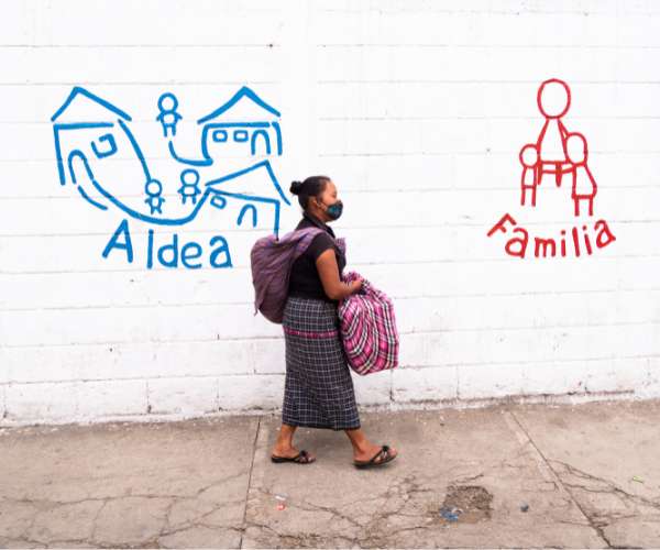 A woman walking on a street in Guatemala with a baby wrapped in a blanket on her back while holding a bag full of supplies